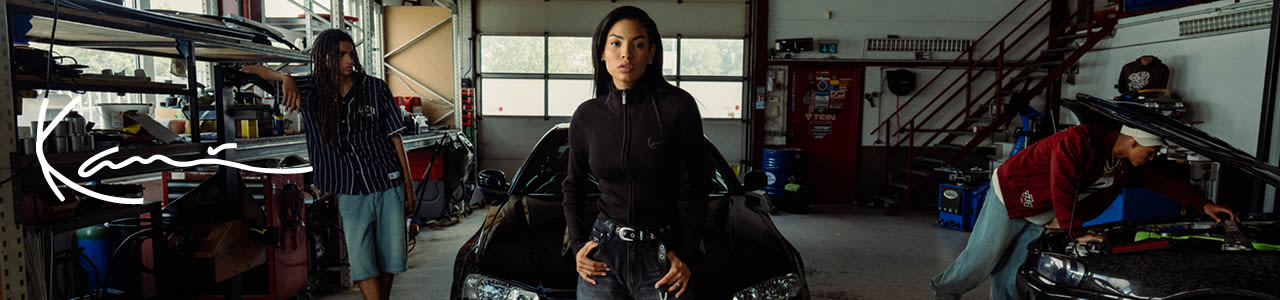 Dual gender models wearing Karl Kani new collection and hanging out in a garage with some cars in the background. Front row: Female model wearing a a black longsleeves turtle neck shirt. In the background, two male models wearing jorts and a blue tshirt, and long jeans and a red college jacket. 