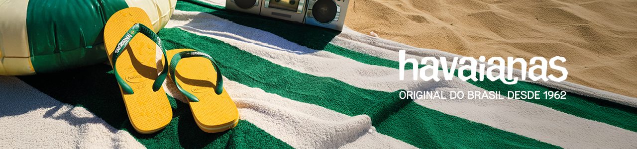 Havaianas yellow and green slides lying on a white and green beach towel. A music radio and sand are in the background