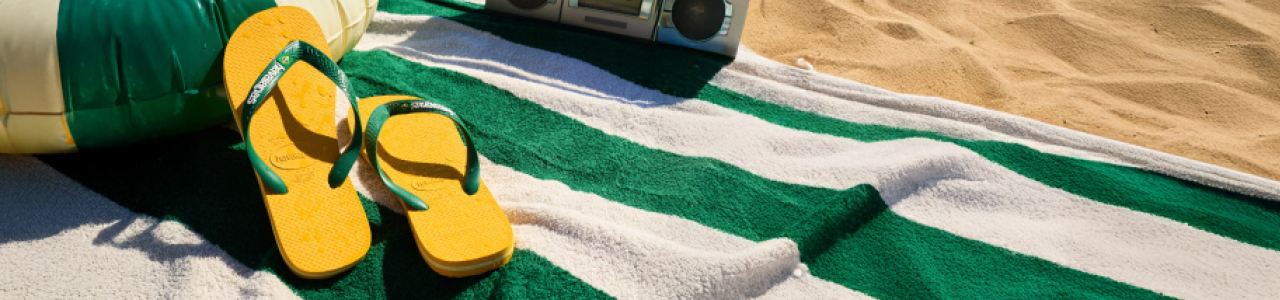 A pair of yellow and green flip-flops resting on a green-and-white striped beach towel, with a volleyball, a boombox, and sand in the background, suggesting a sunny beach scene.