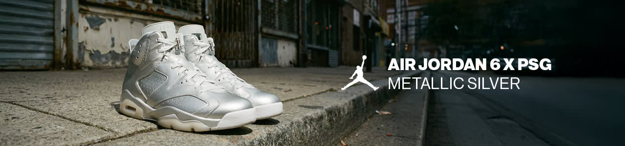 A pair of high-top Air Jordan 6 x PSG sneakers in 'Metallic Silver' featuring a shimmering silver leather upper, white laces, and translucent soles. The shoes are pictured on a city sidewalk against an urban backdrop, accompanied by a white Jumpman logo and text identifying the model.