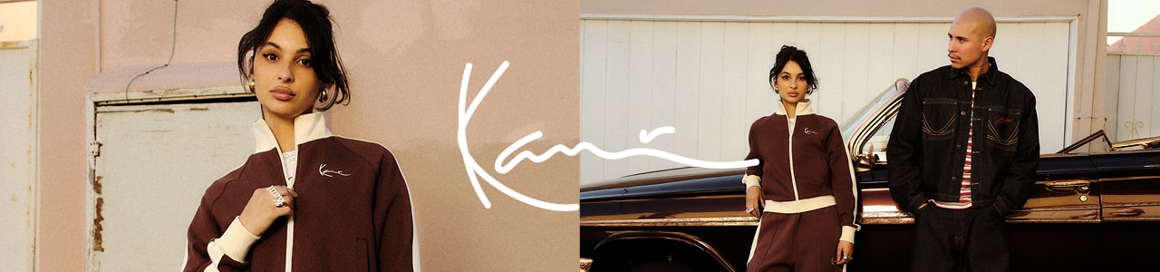 A wide fashion banner for Karl Kani featuring two models in streetwear. On the left, a close-up of a woman wearing a dark burgundy track jacket with white trim and a high collar. In the center, a large white "Kani" signature logo overlays the scene.  On the right, the same woman stands in a full matching tracksuit next to a man wearing a dark denim jacket with yellow contrast stitching and a striped shirt. They are posing in front of a classic, dark-colored car against a light pink building and a white garage door. The lighting is warm and golden, suggesting a sunset or "golden hour" aesthetic.