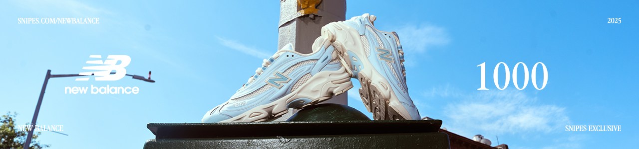 New Balance Sneakers light blue and white at the center of the image, with a blue sky and some beige old building in the background. New Balance 1000. Snipes Exclusive. 2025. snipes.com/newbalance