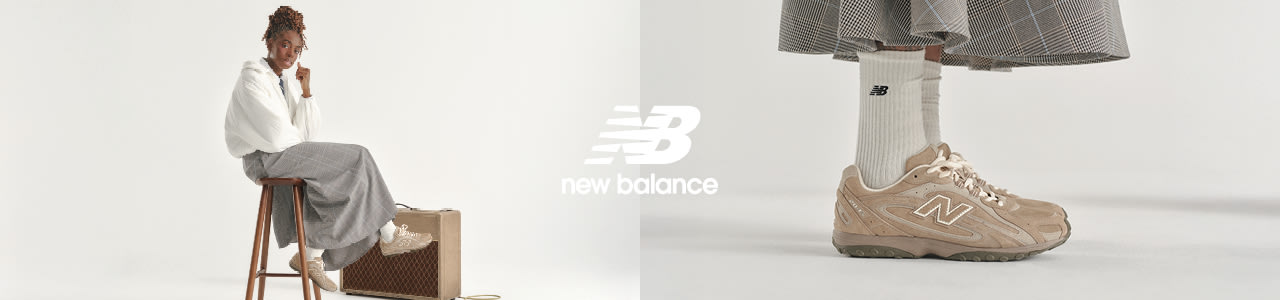 An eye-level, side-profile shot focuses on a person's lower legs and feet. They wear a tan New Balance sneaker with a large, white "N" logo on the side and cream-colored laces. Their white, ribbed crew socks feature a small, black New Balance logo. Above the socks, the bottom of a long, flared, brown and grey plaid skirt is visible. A white New Balance logo is in the top left corner. The background is a solid, light grey.