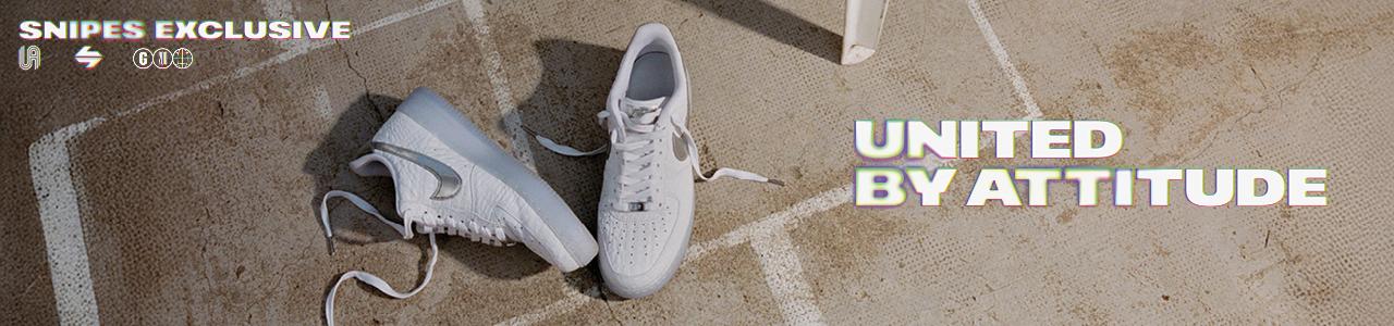 Top-down shot of a pair of white Nike Air Force 1 sneakers resting on a sunlit, textured concrete ground marked with white lines. The shoes feature a crinkled leather upper, metallic silver Swooshes, and translucent icy blue outsoles. One shoe lies on its side while the other stands upright. The top-left corner features the text "UNITED BY ATTITUDE" in a bold, outlined font, and the right side includes a vertical "SNIPES EXCLUSIVE" logo.