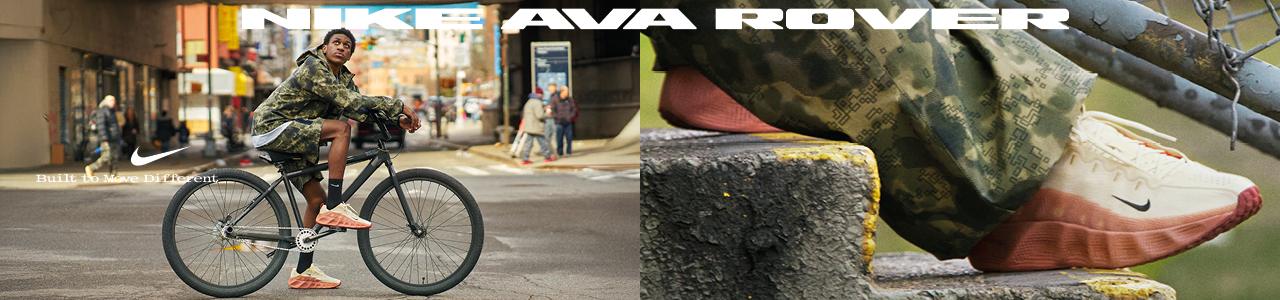 A young man wearing a camouflage jacket and shorts sits on a black bicycle in an urban setting. He is wearing Nike AVA Rover sneakers, which are prominently featured in the image. The shoes are light beige with a bold, thick coral-orange sole. Text on the image reads "NIKE AVA ROVER" at the top and "Built to Move Different" near the shoes, emphasizing the footwear's unique design and functionality.