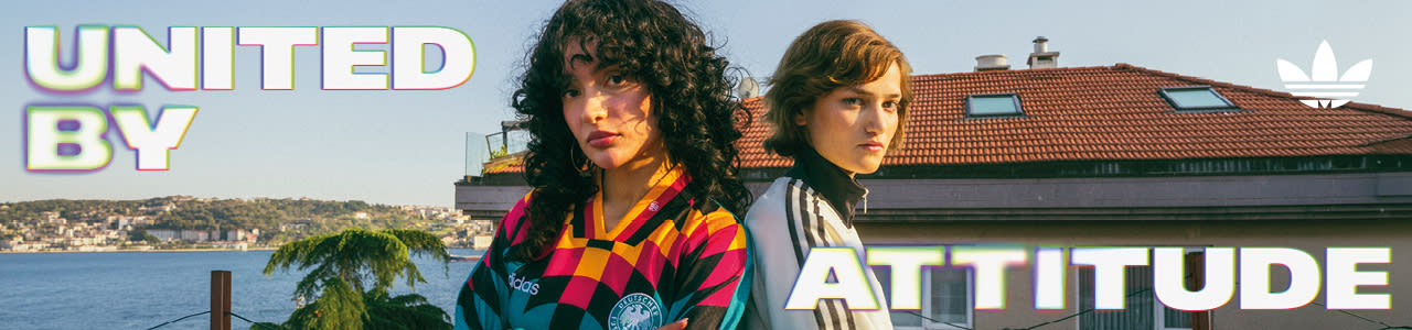 An promotional banner featuring five individuals posing outside a European-style cafe or storefront with a dark green facade. They are all wearing retro-style Adidas athletic apparel, including jerseys and jackets featuring the Germany national football team crest. On the left side of the banner, the text "UNITED BY" is displayed in a bold, white, stylized font with a soft neon glow. On the right side, the text "ATTITUDE" is written in the same font style, alongside a white Adidas Originals trefoil logo in the top right corner. The atmosphere is casual and relaxed, with the individuals sitting and standing around outdoor tables.