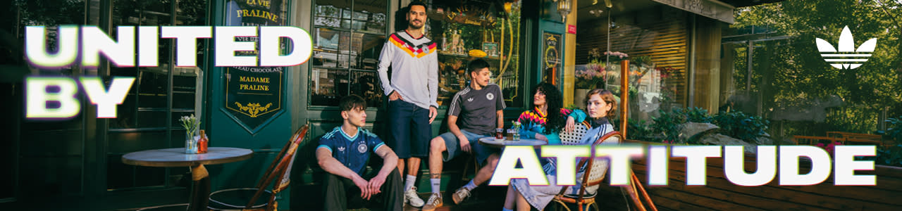 An promotional banner featuring five individuals posing outside a European-style cafe or storefront with a dark green facade. They are all wearing retro-style Adidas athletic apparel, including jerseys and jackets featuring the Germany national football team crest. On the left side of the banner, the text "UNITED BY" is displayed in a bold, white, stylized font with a soft neon glow. On the right side, the text "ATTITUDE" is written in the same font style, alongside a white Adidas Originals trefoil logo in the top right corner. The atmosphere is casual and relaxed, with the individuals sitting and standing around outdoor tables.