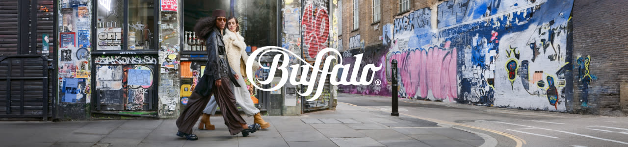 A panoramic low-angle shot of two models, one Black and one white, walking in stride along an urban sidewalk in Shoreditch, London. They are surrounded by walls and metal security shutters completely covered in various styles and colors of graffiti, including large pink letters and diverse sticker bombed sections. The Black model in the foreground has curly dark hair and is wearing sunglasses, a dark overcoat, wide-legged dark trousers, and dark-heeled boots. The white model behind her has light hair and is wearing a light fluffy coat, lighter trousers, and tan chunky-heeled platform boots. A large, prominent white script logo "Buffalo" is superimposed in the center of the image. The pavement is stone slab, and a single black bollard stands to the right.