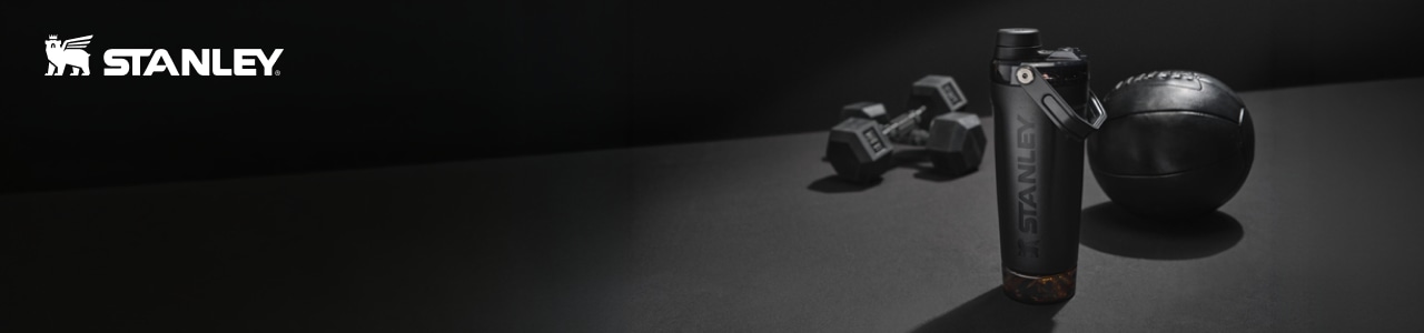 A black Stanley water bottle sits on a dark gym surface beside a medicine ball and two hex dumbbells, with the white Stanley logo displayed on the left; the lighting highlights the matte textures for a sleek, athletic look.