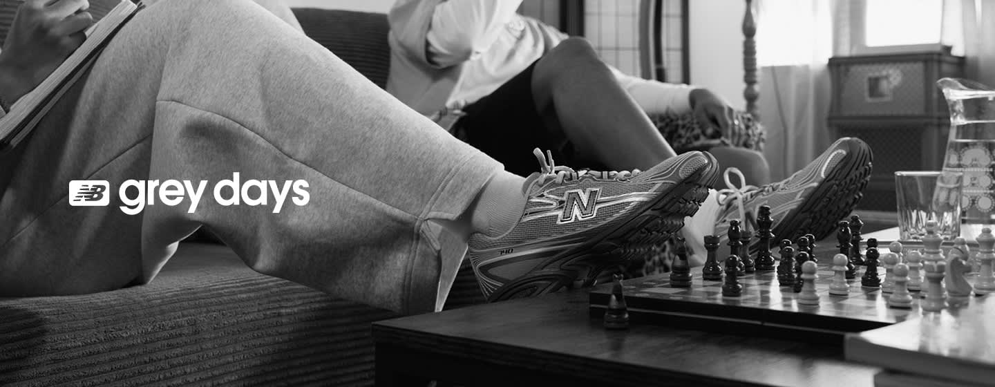 A black and white lifestyle photograph for New Balance's "Grey Days" campaign. The image features two people lounging on a couch; one person is wearing grey sweatpants and a pair of New Balance sneakers with their feet propped up on a wooden coffee table next to a chess set. The text "NB grey days" is overlaid in white on the left side of the frame