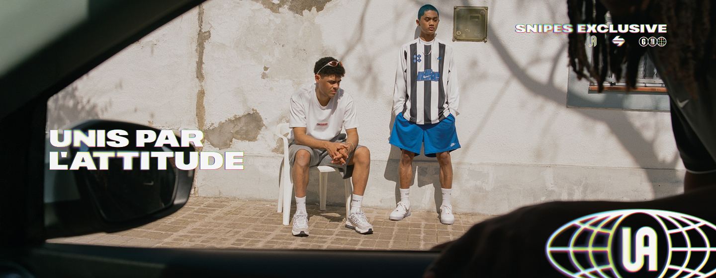 An aerial, slightly angled shot shows a pair of white Nike Air Force 1 sneakers resting on a sunlit, textured concrete ground. The shoes feature metallic silver Swooshes and translucent outsoles.  In the upper-left corner, the text "SNIPES EXCLUSIVE" appears above three stylized logos. In the bottom-right corner, the French slogan "UNIS PAR L'ATTITUDE" (United by Attitude) is written in a bold, white font with a colorful, glitch-effect outline. The scene is bright with soft shadows cast by the sneakers.