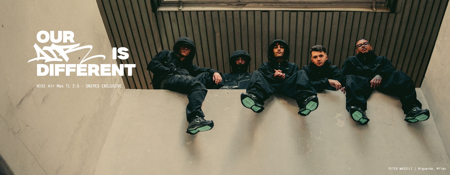 A young man in a black hooded tracksuit sits outdoors in front of tall apartment buildings, holding up a pair of black Nike Air Max TL 2.5 sneakers with green soles. The ad features bold white text reading “OUR AIR IS DIFFERENT” along with “NIKE Air Max TL 2.5 – SNIPES EXCLUSIVE.” At the bottom right