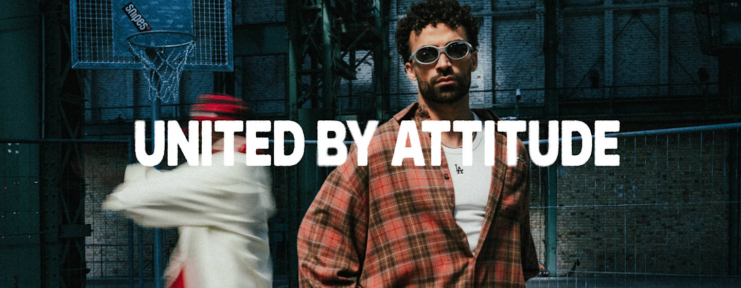 Majid is standing in front of the camera at the center of the image, wearing a white tanktop and a plaid shirt. 2 dancers are dancing in the background and the movement is blurred. United by Attitude claim is written at the center of the image in white font. 