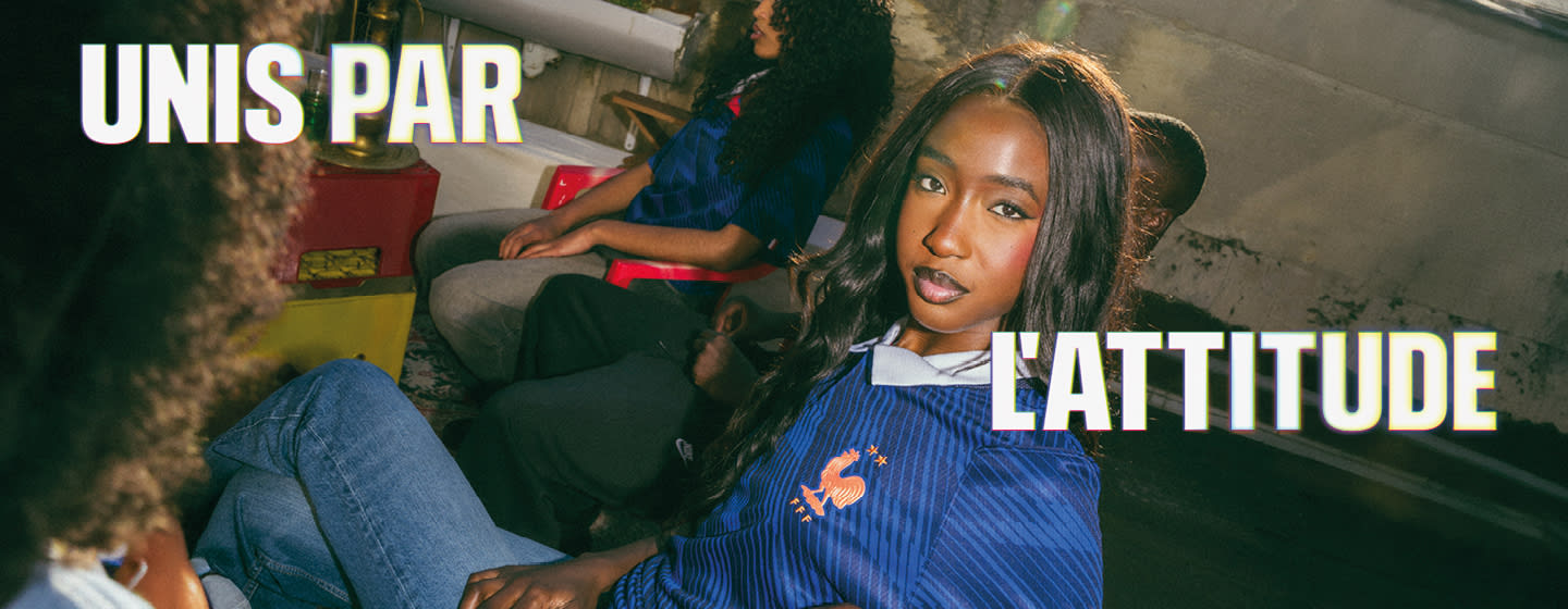 Wide banner featuring three smiling young adults on an urban rooftop, dressed in Nike France soccer apparel. Overlaid on the left and right in large, bold white text with a subtle yellow glow is the French phrase "UNIS PAR L'ATTITUDE". On the left, a man sits in a light-colored jacket with black and red accents. In the center and right, two women sit together wearing blue patterned France national team jerseys with the FFF rooster crest.