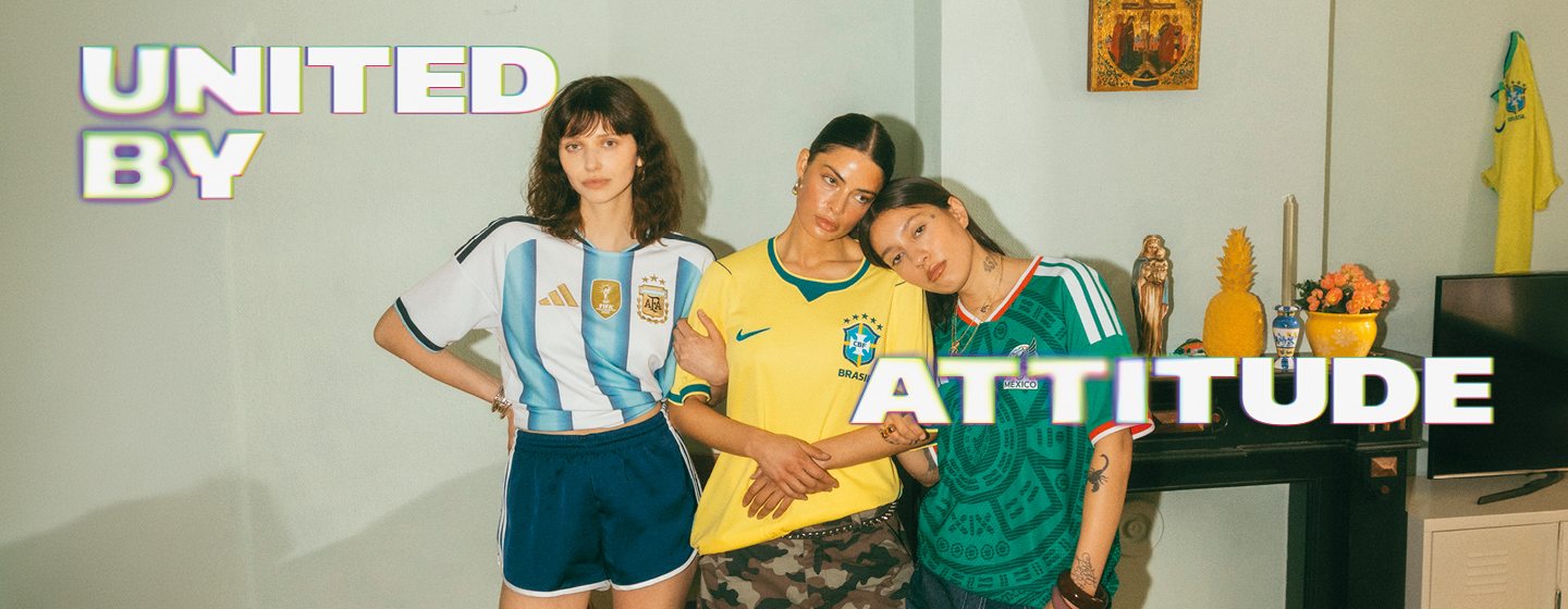 A promotional banner featuring five young individuals hanging out during a casual outdoor gathering in a backyard or patio setting. One person sits near a small charcoal grill with food cooking on it, while the others are seated around a patio table with food and drinks. They are all wearing different national football team jerseys, including France, Italy, Brazil, and Argentina. Large, stylized text overlays the image, with "UNITED BY" in the top-left corner and "ATTITUDE" across the bottom right, both featuring a bold white font with a soft neon glow.