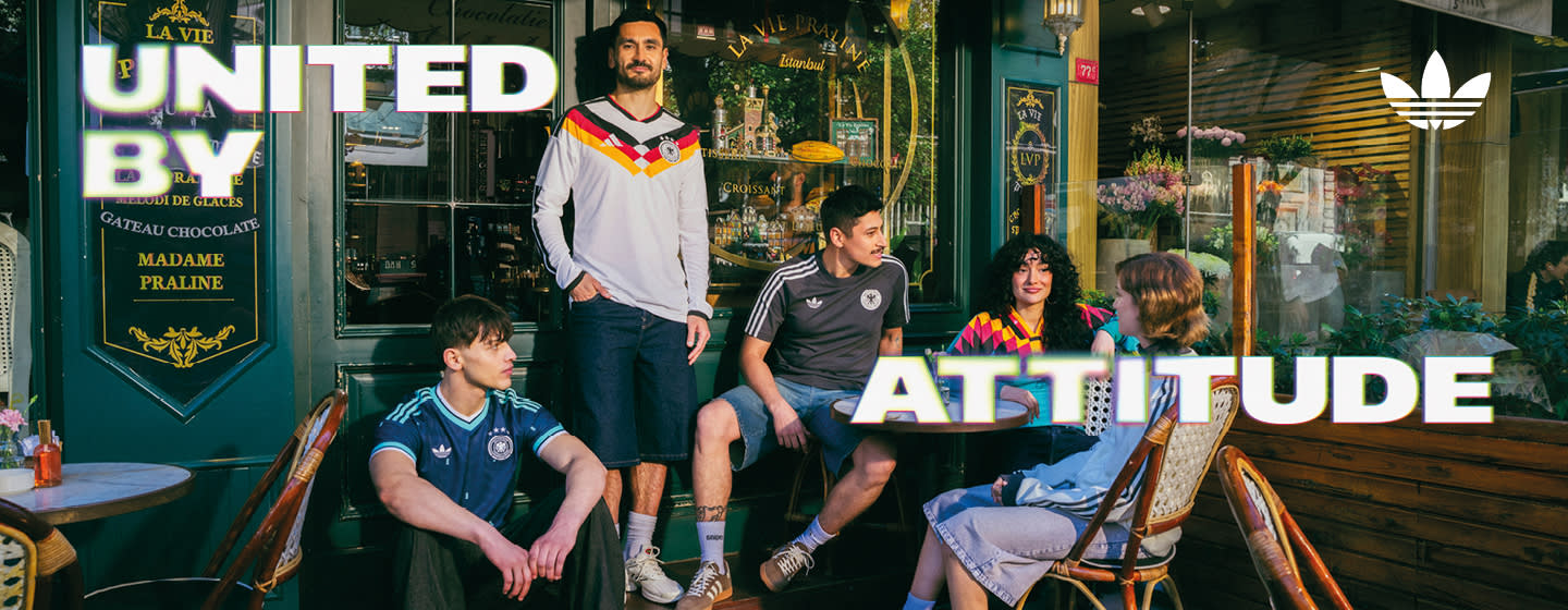 An promotional banner featuring five individuals posing outside a European-style cafe or storefront with a dark green facade. They are all wearing retro-style Adidas athletic apparel, including jerseys and jackets featuring the Germany national football team crest. On the left side of the banner, the text "UNITED BY" is displayed in a bold, white, stylized font with a soft neon glow. On the right side, the text "ATTITUDE" is written in the same font style, alongside a white Adidas Originals trefoil logo in the top right corner. The atmosphere is casual and relaxed, with the individuals sitting and standing around outdoor tables.