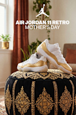 A promotional image for the "Air Jordan 11 Retro Mother's Day" sneakers. A pair of white low-top sneakers with a metallic gold patent leather mudguard are displayed on a black velvet ottoman with intricate gold embroidery. The background features a cozy living room setting with potted plants, a warm orange curtain, and a wooden armchair with a throw pillow. The Jumpman logo and the text "AIR JORDAN 11 RETRO MOTHER'S DAY" are overlaid in white at the top of the image.