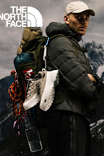 A man is depicted rappelling with arms crossed next to a giant mountain poster, against a brick wall. This is a print ad by The North Face.