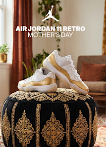 A promotional image for the "Air Jordan 11 Retro Mother's Day" sneakers. A pair of white low-top sneakers with a metallic gold patent leather mudguard are displayed on a black velvet ottoman with intricate gold embroidery. The background features a cozy living room setting with potted plants, a warm orange curtain, and a wooden armchair with a throw pillow. The Jumpman logo and the text "AIR JORDAN 11 RETRO MOTHER'S DAY" are overlaid in white at the top of the image.