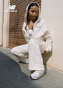 Woman wearing a cream-colored hoodie and sweatpants, crouching against a wall with braided hair, sporting white and silver New Balance sneakers. The New Balance logo appears in the top left corner.