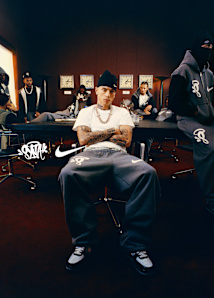 A panoramic promotional banner for a Nike collaboration featuring British rapper Central Cee. The left side is a portrait of Central Cee wearing a dark grey Nike zip-up hoodie with the hood up and rimless glasses, positioned next to a white Nike swoosh and a "Syna" graffiti-style logo. The right side shows a wide shot of Central Cee sitting at a conference table in a moody, corporate setting, surrounded by other men. He is wearing a white t-shirt, grey Nike sweatpants, a black beanie, and silver jewelry. The Nike swoosh is prominently displayed on the apparel and the wall.