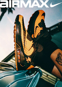 A split-screen Nike Air Max advertisement banner set outdoors under a clear sky. On the left side, two young men are looking toward the camera; one stands in the background wearing a black graphic t-shirt and camouflage pants while holding a flag pole, and the other sits in the foreground wearing a dark checkered short-sleeve button-up shirt featuring a small skull and crossbones logo. The word "AIRMAX" with the Nike swoosh is displayed in large white text across the top left. On the right side, a close-up, angled shot shows a black and mustard-yellow Nike Air Max sneaker with a visible air bubble in the heel and a small orange skull and crossbones graphic near the ankle.