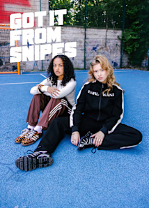 Two young women are sitting on a bright blue outdoor sports court in front of a fence and graffiti-covered wall. The woman on the left has curly hair, wears a white long-sleeve top, brown Adidas track pants with white stripes, and leopard-print Adidas sneakers. The woman on the right has wavy blonde hair, wears a black Karl Kani tracksuit with white side stripes, and black-and-white striped sneakers. Large bold white text above them reads, “GOT IT FROM SNIPES.”