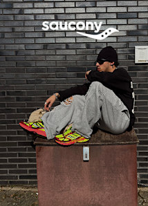 An outdoor lifestyle banner for Saucony featuring a person sitting on a metal railing of a modern, brutalist-style building under a cloudy sky.  On the left, the person is shown in a wide shot wearing a grey hoodie, dark trousers, a beanie, and vibrant purple and lime green Saucony sneakers. The center displays the Saucony logo in white against a grey, overcast sky. On the right, a close-up focuses on the person's feet dangling, highlighting the detailed design of the Saucony sneakers, which feature silver overlays and bright yellow soles.