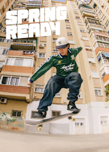 Man performing a skateboard trick, wearing Adidas shoes, a green long-sleeve, jeans, and a Yankees cap, with a beige building in the background.