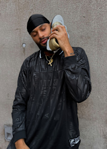 Male model wearing all black football inspired streetwear apparel and white and gold Nike Total 90 shoes, standing outside against a concrete wall