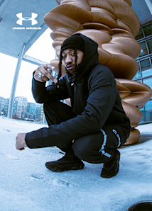 A person dressed in a black Under Armour "Unstoppable" hooded jacket, beanie, and matching joggers crouches in a snowy outdoor setting. The scene features a futuristic architectural backdrop with a large, bronze-colored abstract sculpture. One view highlights a close-up of the person tying the laces of a black Under Armour athletic shoe against the snow. The Under Armour logo is prominently displayed.