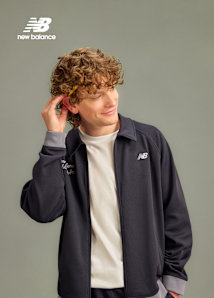 A medium-length portrait of a smiling person with curly light brown hair wearing a dark navy New Balance track jacket over a white t-shirt. Their right hand is reaching behind their ear. In the upper left corner, the New Balance logo is visible in white. The background is a flat, medium grey.