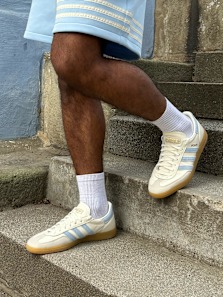 Person wearing cream Adidas sneakers with light blue stripes, white socks, and light blue shorts stands on concrete steps with a textured urban background.