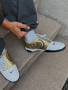 Male model wearing all black football inspired streetwear apparel and white and gold Nike Total 90 shoes, standing outside against a concrete wall