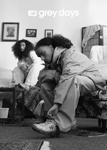 A black and white lifestyle photograph for New Balance's "Grey Days" campaign. The image features two people lounging on a couch; one person is wearing grey sweatpants and a pair of New Balance sneakers with their feet propped up on a wooden coffee table next to a chess set. The text "NB grey days" is overlaid in white on the left side of the frame