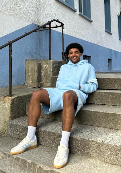 Guy sitting outside in the stairs with a light blue adidas hoodie and shorts smiling 