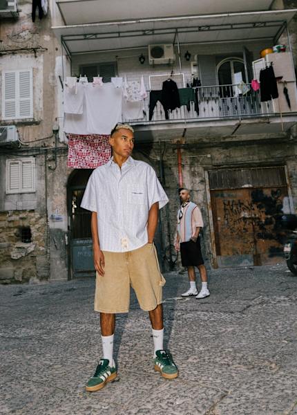 Two young men pose in a gritty, urban courtyard with weathered buildings and laundry hanging from a balcony. The man in the foreground wears a light blue striped short-sleeve shirt, tan corduroy shorts, white socks, and green Adidas sneakers. The man in the background wears a retro-style striped shirt, black shorts, and white Adidas sneakers. The setting features cracked stone pavement, air conditioning units, graffiti, and a strong street-style aesthetic.