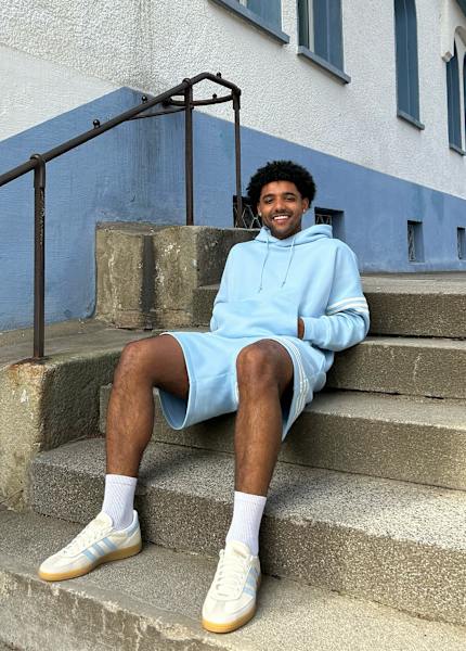 Guy sitting outside in the stairs with a light blue adidas hoodie and shorts smiling 