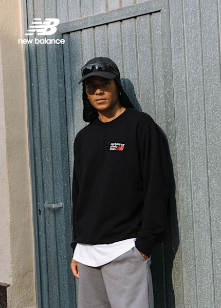 A man wearing a black New Balance sweatshirt, gray shorts, white socks, and gray New Balance sneakers sits casually on the ground against a wall. He also wears a black cap with attached sunglasses. The New Balance logo is displayed in the top left corner.