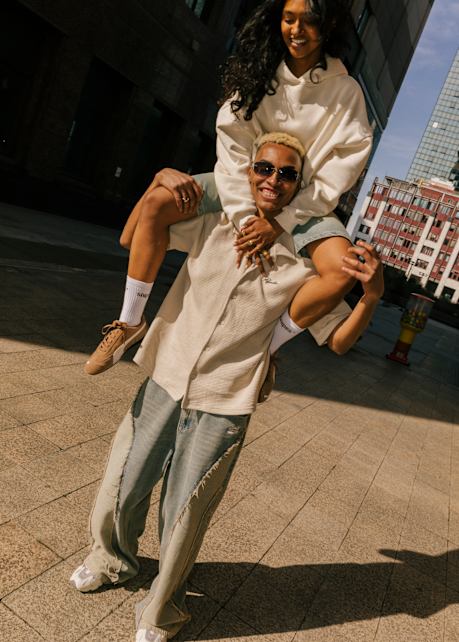 male and female model in urban environment wearing summer streetwear. The woman is sitting the man's neck. Both are smiling.