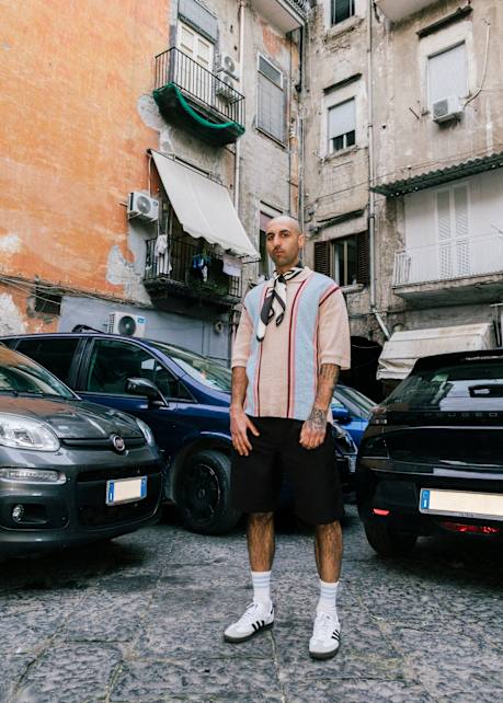 A man stands confidently in a narrow courtyard between parked cars, framed by weathered apartment buildings with peeling paint and balconies. He wears a beige short-sleeve knit shirt with red and blue vertical stripes, dark shorts, white socks, and white Adidas sneakers. A scarf is loosely tied around his neck, and his tattoos are visible on his arm. The setting evokes a gritty, urban European atmosphere.
