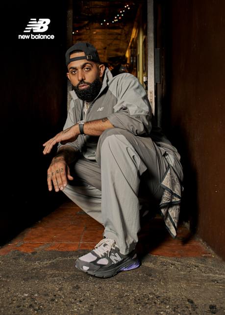 A man with a beard, tattoos, and a black baseball cap wears a grey New Balance tracksuit and grey 990v3 sneakers with purple accents while posing against a red wall and in a dark hallway; a supporting image shows a group of people wearing the same sneakers.