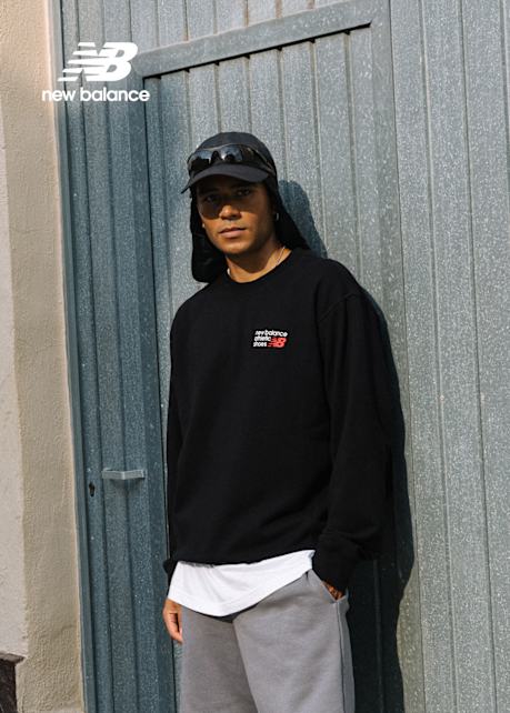 A man wearing a black New Balance sweatshirt, gray shorts, white socks, and gray New Balance sneakers sits casually on the ground against a wall. He also wears a black cap with attached sunglasses. The New Balance logo is displayed in the top left corner.