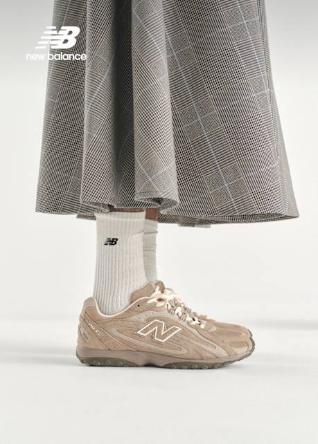 An eye-level, side-profile shot focuses on a person's lower legs and feet. They wear a tan New Balance sneaker with a large, white "N" logo on the side and cream-colored laces. Their white, ribbed crew socks feature a small, black New Balance logo. Above the socks, the bottom of a long, flared, brown and grey plaid skirt is visible. A white New Balance logo is in the top left corner. The background is a solid, light grey.