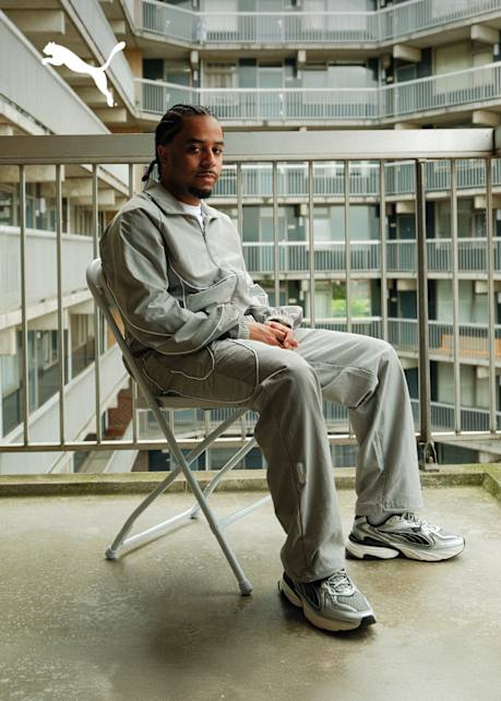 Close-up of a person putting on a black and gray Puma athletic shoe, showing the signature Puma stripe logo and a thick midsole, set in an outdoor environment.