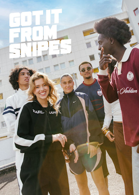 A group of six young adults is standing outdoors in front of a modern white apartment building with yellow balcony accents, smiling and chatting. They are wearing casual streetwear, including branded tracksuits, sweatshirts, and sports jerseys. The text “GOT IT FROM SNIPES” is displayed in bold white block letters at the top left of the image. Sunlight flares slightly across the bottom of the photo, adding a warm and energetic feel.