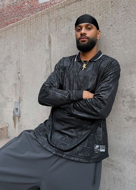 Male model wearing all black football inspired streetwear apparel and white and gold Nike Total 90 shoes, standing outside against a concrete wall