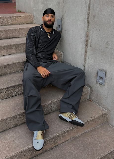 Male model wearing all black football inspired streetwear apparel and white and gold Nike Total 90 shoes, standing outside against a concrete wall