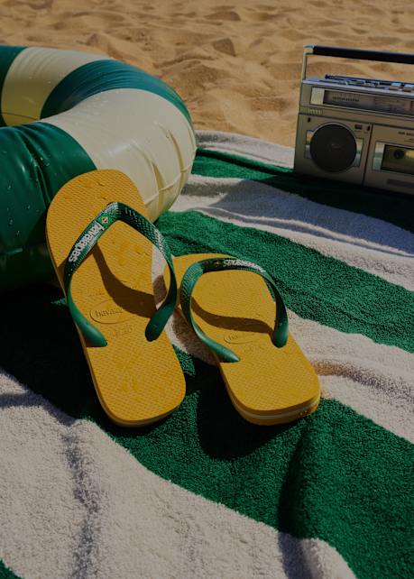 A pair of yellow and green flip-flops resting on a green-and-white striped beach towel, with a volleyball, a boombox, and sand in the background, suggesting a sunny beach scene.