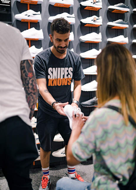 Man standing and presenting white sneaker to seated person in SNIPES store.